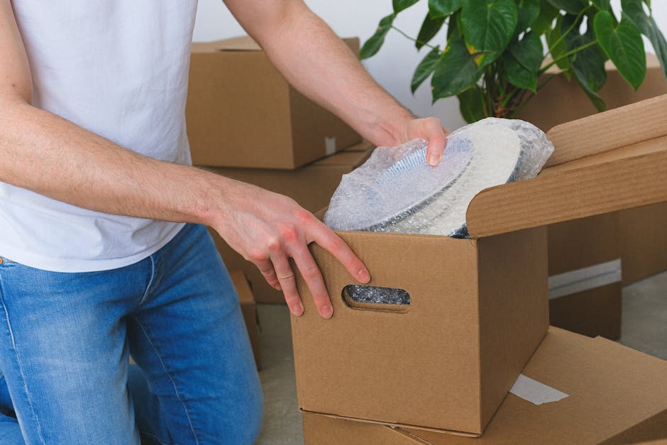 A person wearing a white t-shirt and blue jeans is seen handling a cardboard box during packing in a home environment. The individual is carefully placing a fragile, circular glass item wrapped in bubble wrap into the open top of the box. Several other cardboard boxes are stacked nearby, some closed and others open, indicating an ongoing packing process. The setting features a light-colored room with natural lighting, and a large green leafy plant can be seen in the background, suggesting a domestic interior space preparing for a home relocation. This scene illustrates the careful handling and packing of delicate items as part of furniture transport or moving logistics, aligning with services provided by Removal Companies Surrey during house removals or moving services.