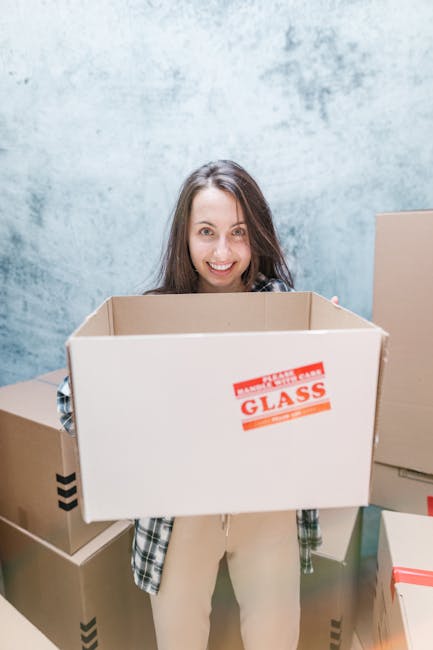 A smiling woman with long brown hair, standing inside a room with a textured light blue wall, holding an open cardboard box labeled 'FRAGILE GLASS' in red text. The box appears to be part of a house relocation or furniture transport process. Surrounding her are additional packed cardboard boxes, some with black tape sealing, and others with plain brown surfaces. The scene suggests packing and moving activities overseen by Removal Companies Surrey, with equipment like packing materials possibly used for safe transport of fragile items. The lighting is bright and even, highlighting the careful handling involved in the loading and moving process relevant to house removals and property relocation tasks.