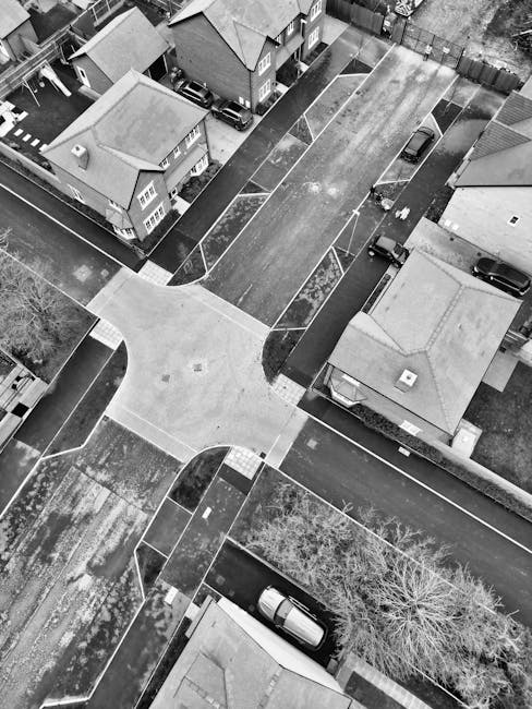 An aerial view in black and white of a residential street with multiple houses and driveways. A large van from Removal Companies Surrey is parked on the road, positioned near the entrance of a drivewa