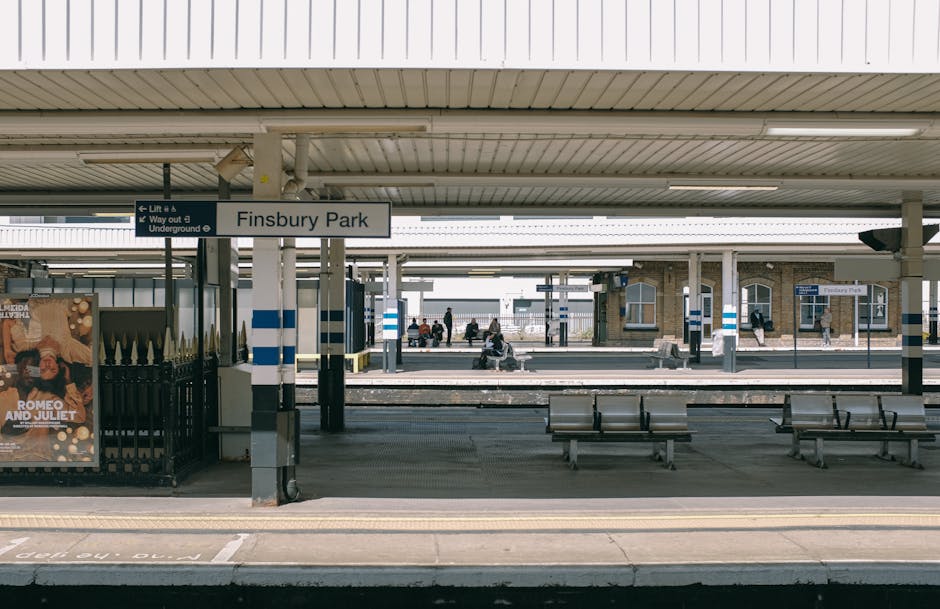 A view of Finsbury Park train station platform with several benches for passengers, surrounded by a covered roof with white and blue stripes, and a sign indicating direction to lift and underground station. On the platform, there are a few commuters seated and standing, some carrying luggage or bags, with a poster advertising 'Romeo and Juliet' visible on a fence to the left. The station environment includes metal fencing, signposts, and a brick building in the distance. This setting illustrates the typical scene during a home relocation or furniture transport process, where belongings are likely being prepared for loading into a vehicle. The communal area, with its open space and seating, is used for waiting or organizing transport logistics before moving items into a removal vehicle, such as a van or truck, which might be parked nearby or in the loading area. The image emphasizes the importance of efficient moving arrangements at transportation hubs during a professional removals service provided by Removal Companies Surrey.